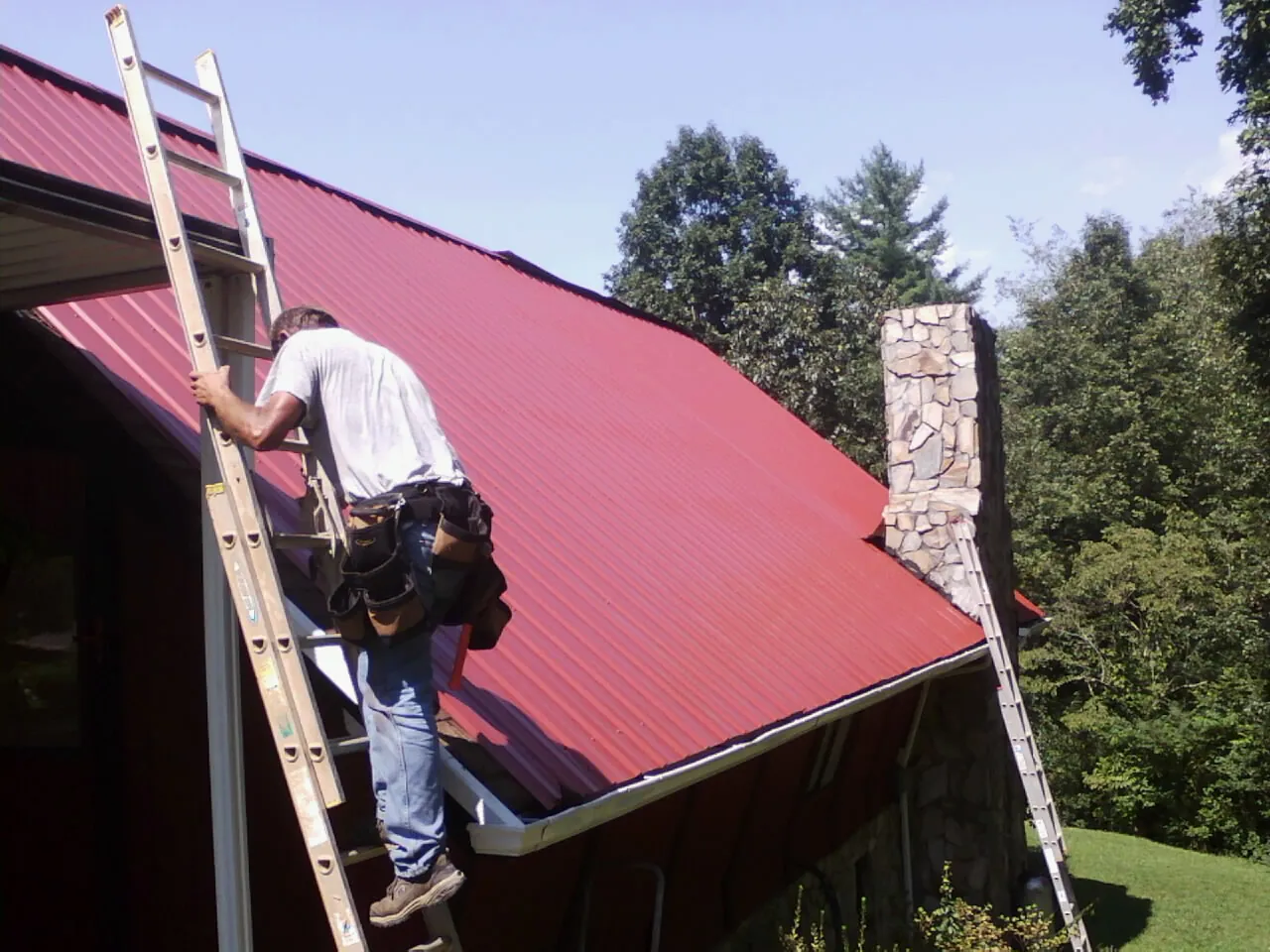 Roofer on a ladder beside a red metal roof and stone chimney — Soffit & Fascia Repair by Roofing Brothers in Falls Church